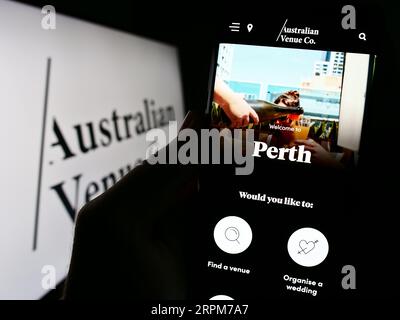 Person holding cellphone with website of company Australian Venue Co. (AVC) on screen in front of logo. Focus on center of phone display. Stock Photo