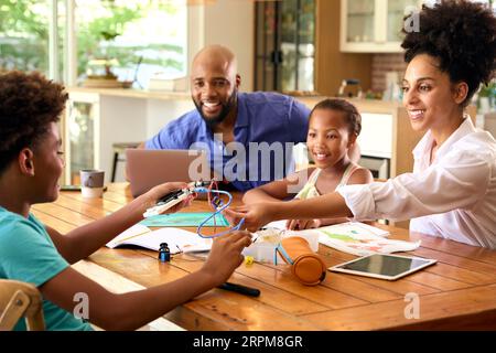 Family Around Table At Home Using Laptop With Parents Helping Children With Science Homework Stock Photo