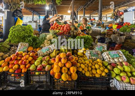 A market trader in a black top stretches both arms over a heap of tomatoes at her fruit and vegetable stall in the covered farmers' market on Chavchavadze Avenue in Telavi, Georgia. Peaches, apples, lemons, bananas and green-and-red fruit, possibly mangoes, lie in the foreground, along with button mushrooms in trays. Behind them are piled potatoes, lettuce, dill, spring onions, purple basil and cherry tomatoes. On the right stands a woman in a colourful dress holding a purse. Vegetable stalls line up in the background beneath a wooden ceiling with steel girders. A curved roof covers the main s Stock Photo