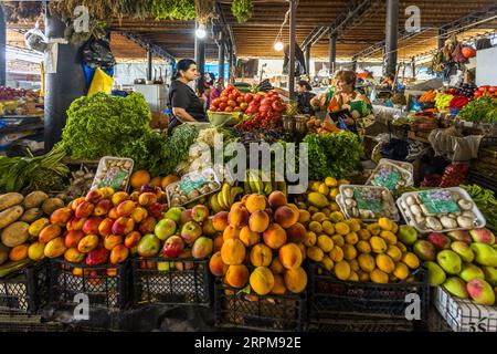 A fruit and vegetable stall in the covered farmers' market on Chavchavadze Avenue in Telavi, Georgia, sells peaches, apples, lemons, bananas and green-and-red fruit, possibly mangoes. Button mushrooms lie in film-wrapped trays. Behind them are piled potatoes, lettuce, dill, spring onions, purple basil, tomatoes and cherry tomatoes. A market trader in a black top stands behind the display. On the right a woman in a colourful dress holds a purse. Bundles of dried herbs hang beneath the wooden ceiling with its steel girders. A curved roof covers the main section of the market, with stalls arrange Stock Photo