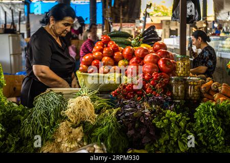 A vegetable and herb stall at the covered farmers’ market in Telavi, Kakheti: a market trader inspects the produce on offer, including large beef tomatoes, cherry tomatoes on the vine, purple basil (regan), spring onions, dill, carrots and pickled olives in jars. Purple basil is a common herb in Georgian cuisine, added to soups, salads and the festive supra. Ilia Chavchavadze Avenue, Telavi Municipality, Kakheti, 2200, Georgia Stock Photo