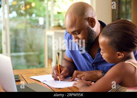 Father And Daughter Around Table At Home Using Laptop For Homework Stock Photo