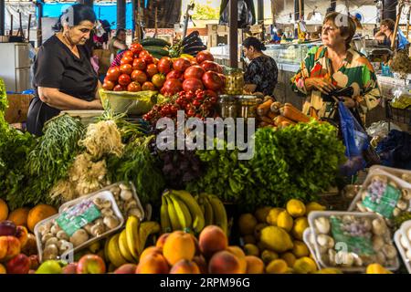 A market trader in a black top looks down at the display of her fruit and vegetable stall in the covered farmers' market on Chavchavadze Avenue in Telavi, Georgia. Button mushrooms in trays, bananas, oranges, peaches, lemons and apples lie in the foreground. Behind them are piled tomatoes, cherry tomatoes on the vine, cucumbers, aubergines, purple basil, dill and spring onions. On the right stands a woman with glasses and a colourful dress, holding an open purse and a blue plastic bag. A curved roof covers the main section of the market, with stalls arranged in parallel rows. The town is the a Stock Photo