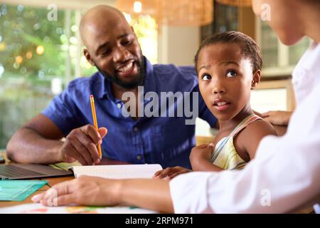 Family Around Table At Home Using Laptop With Parents Helping Daughter With Homework Stock Photo