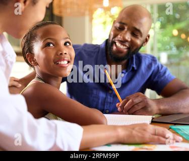 Family Around Table At Home Using Laptop With Parents Helping Daughter With Homework Stock Photo
