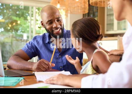 Family Around Table At Home Using Laptop With Parents Helping Daughter With Homework Stock Photo