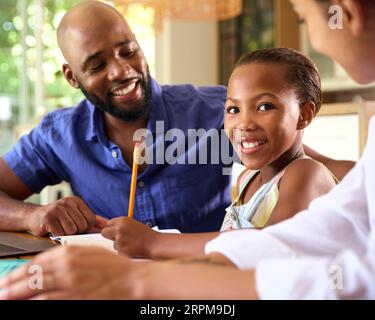 Family Around Table At Home Using Laptop With Parents Helping Daughter With Homework Stock Photo