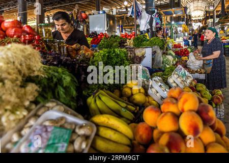 Peaches, bananas and yellow fruit, possibly lemons, lie at the front of a fruit and vegetable stall in the covered farmers' market on Chavchavadze Avenue in Telavi, Georgia. On the left lie fresh garlic and button mushrooms in trays. Behind them stand bunches of green and purple herbs, among them dill. A market trader in a black top stands behind the herbs, next to tomatoes, carrots and jars of pickled vegetables. On the right, an elderly woman in a green headscarf holds a piece of fruit, with apples in front of her. Stalls with tomatoes, cabbage and cauliflower line the background. A curved r Stock Photo
