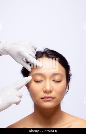 Hands of beautician in white surgical gloves touching face of asian woman with dark hair, copy space Stock Photo