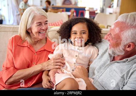 Granddaughter Cuddling With Loving Grandparents On Sofa At Home With Parents In Background Stock Photo