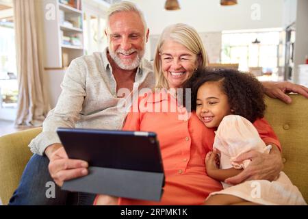 Granddaughter And Grandparents On Sofa At Home Making Video Call On Digital Tablet Stock Photo