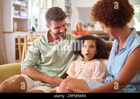 Daughter Sitting With Parents On Sofa At Home Pulling Funny Face With Grandparents In Background Stock Photo