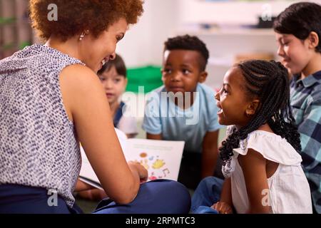 Female Teacher Reads To Multi-Cultural Elementary School Pupils Sitting On Floor In Class At School Stock Photo