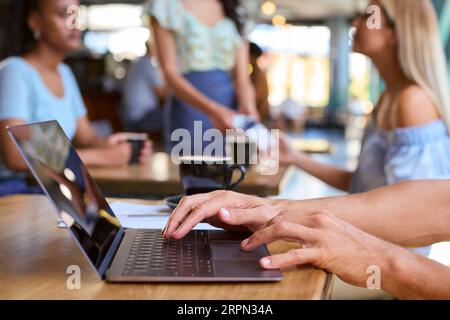 Close Up Of Man Working On Laptop In Foreground With Women Meeting Behind In Busy Coffee Shop Stock Photo