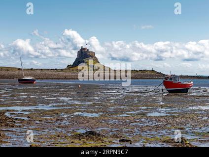 View towards Lindisfarne Castle standing on its rocky crag seen across the muddy harbour at low tide with boats aground on the sand. Stock Photo