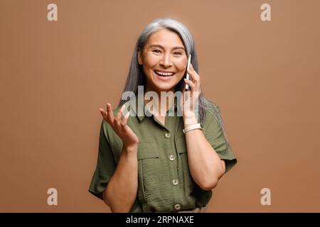 Cheerful happy mature Asian woman talking on smartphone isolated on brown background, 40s lady having pleasant phone conversation, chatting with friend Stock Photo