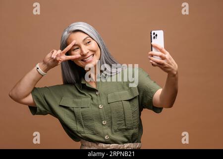 Mature Asian woman wearing casual green shirt with gray long hair standing isolated on brown background, taking selfie photo on smartphone looking camera laughing happy close-up Stock Photo