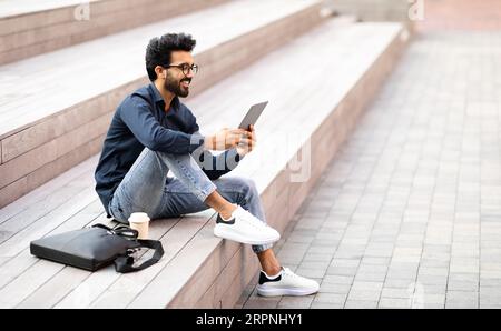 Relaxed indian guy chilling outdoor after work, using digital tablet Stock Photo