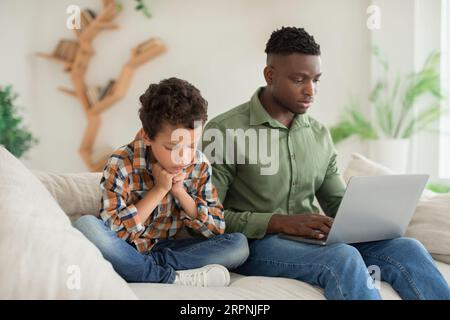 Black Young Father Ignoring Unhappy Little Son Using Laptop Indoor Stock Photo
