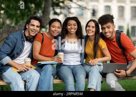 Outdoor Portrait Of Happy Young Multiethnic Students Relaxing On Bench At Campus Stock Photo