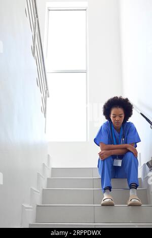 Stressed Female Doctor Or Nurse Wearing Scrubs Sitting On Stairs In Hospital Stock Photo