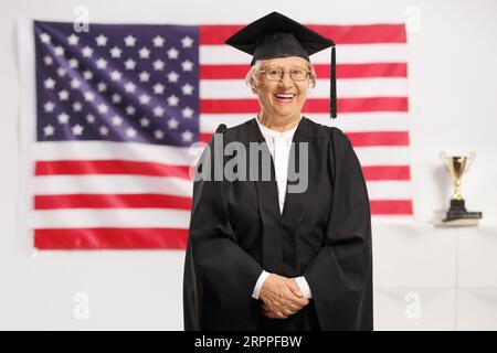 Elderly female wearing a graduation gown and smiling in front of a USA flag Stock Photo