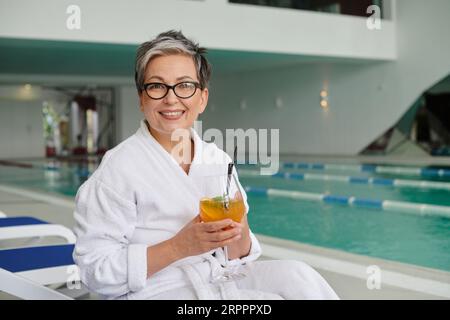 spa center concept, happy mature woman in glasses and robe holding cocktail and sitting on lounger Stock Photo