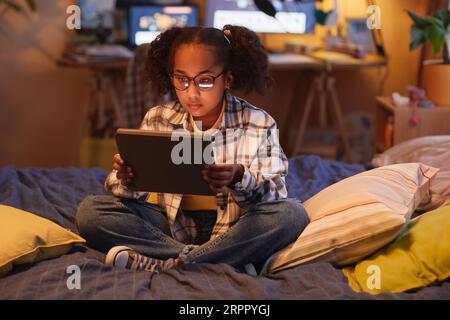 Full length portrait of teen black girl with pigtails using digital tablet while sitting on bed at home in cozy warm light, copy space Stock Photo