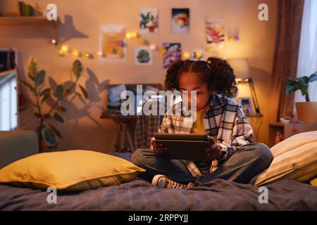 Full length portrait of teenage black girl with pigtails using digital tablet while sitting on bed at home in cozy warm light, copy space Stock Photo