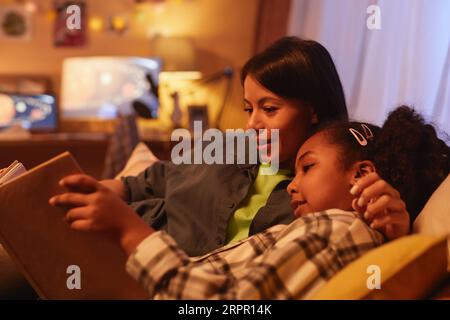 Portrait of young Black mother and daughter lying in bed together and reading book at night Stock Photo