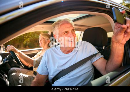 Male Passenger Taking Selfie On Mobile Phone As Two Senior Couple Enjoy Day Trip Out In Car Stock Photo