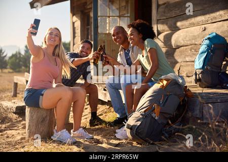 Group Of Friends On Vacation Sitting On Porch Of Countryside Cabin Drinking Beer And Taking Selfie Stock Photo