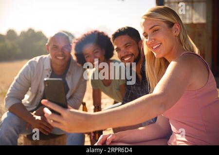 Group Of Friends On Vacation Sitting On Porch Of Countryside Cabin Drinking Beer And Taking Selfie Stock Photo