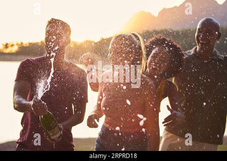 Group Of Friends Celebrating Outdoors By Lake Opening And Spraying Bottle Of Champagne Stock Photo