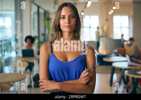Portrait Of Smiling Young Businesswoman Holding Glasses Standing In Busy Office Stock Photo