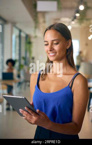 Young Businesswoman Standing In Busy Office Using Digital Tablet Stock Photo
