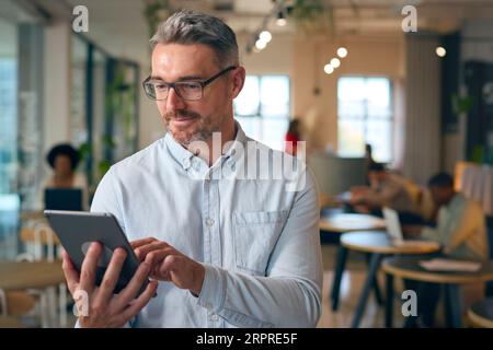 Mature Businessman Standing In Busy Office Using Digital Tablet Stock Photo