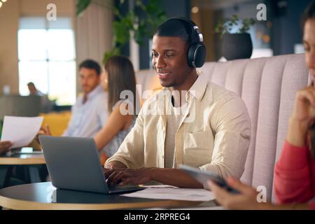 Businessman Wearing Wireless Headphones Working On Laptop In Informal Seating Area Of Modern Office Stock Photo