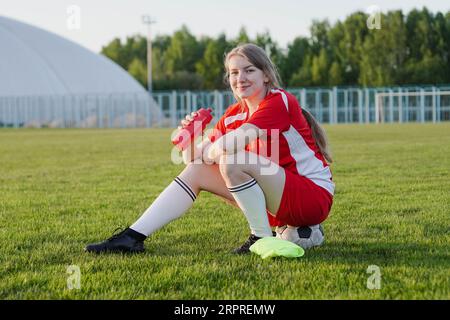Teen girl in red football strip sitting on soccer ball holding bottle of water at sunset, smiling to camera Stock Photo