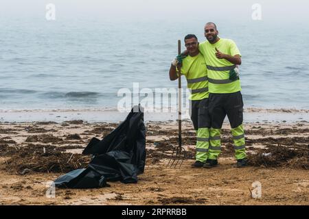 Two young male municipal workers clearing up the beach in Cascais, Portugal pose for a picture smiling with thumbs up gesture Stock Photo