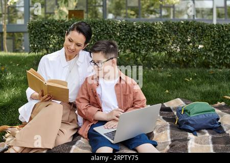 happy middle aged woman reading book near son with down syndrome using laptop in park Stock Photo
