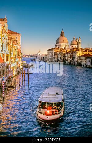 Venice Grand Canal with Vaporetto passenger waterbus water bus regular scheduled transport in foreground and Basilica di Santa Maria della Salute behind at sunset Venice Italy Stock Photo