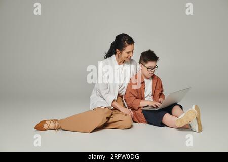 kid with down syndrome sitting near smiling mother and using laptop on grey, full length Stock Photo