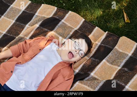 top view of carefree boy with down syndrome, in eyeglasses, sticking out tongue on blanket in park Stock Photo