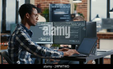 Portrait of african american developer using laptop to write code sitting at desk with multiple screens parsing algorithm in software agency. Coder working on user interface using portable computer. Stock Photo