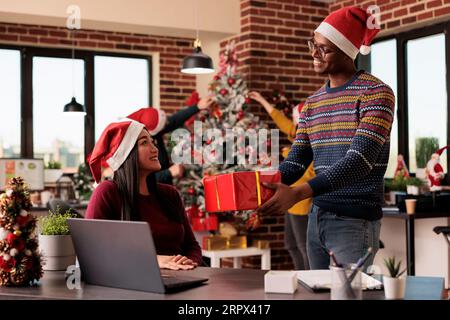 Smiling african american office worker giving cristmas present to asian woman colleague in decorated office. Diverse employees sharing xmas gifts while working on laptop in workspace Stock Photo