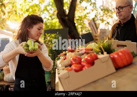 Two farmers vendors putting fruits and veggies on market stand, preparing for new day to sell organic produce. Man and woman wokring at local farmers market, fresh natural food. Stock Photo