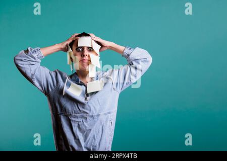 Exhausted unhappy woman writes down memo messages using paper sticky notes, helping her with plenty details about work. Colorful memory reminder on tired depressed young adult, studio shot Stock Photo