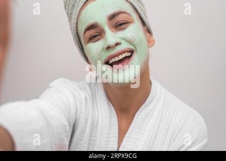 Joyful woman blogger with clay mask on face wearing bathrobe making selfie on white background Stock Photo