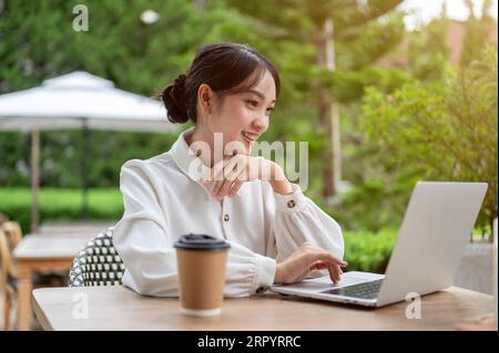 An attractive Asian female freelancer is working remotely at a beautiful cafe's outdoor seating area, using her laptop computer to manage her tasks. U Stock Photo
